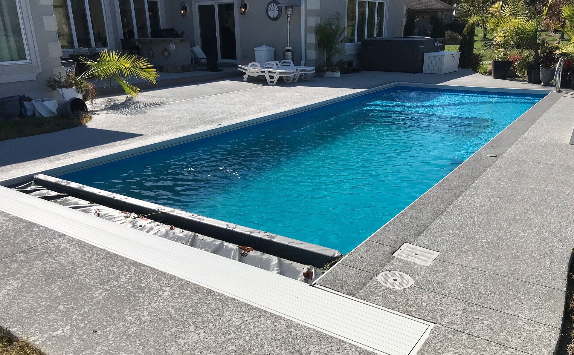 Rectangular swimming pool with blue water surrounded by concrete patio. A house is in the background.