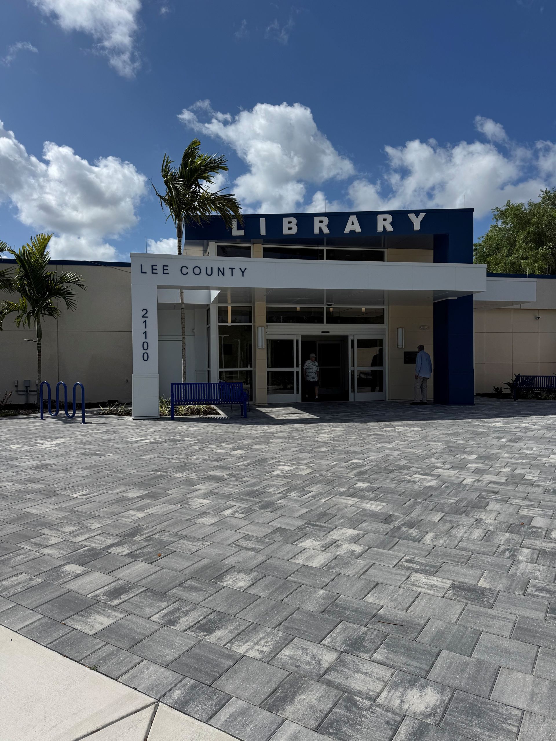 Library entrance with blue sign, white building, and paved courtyard under a bright sky