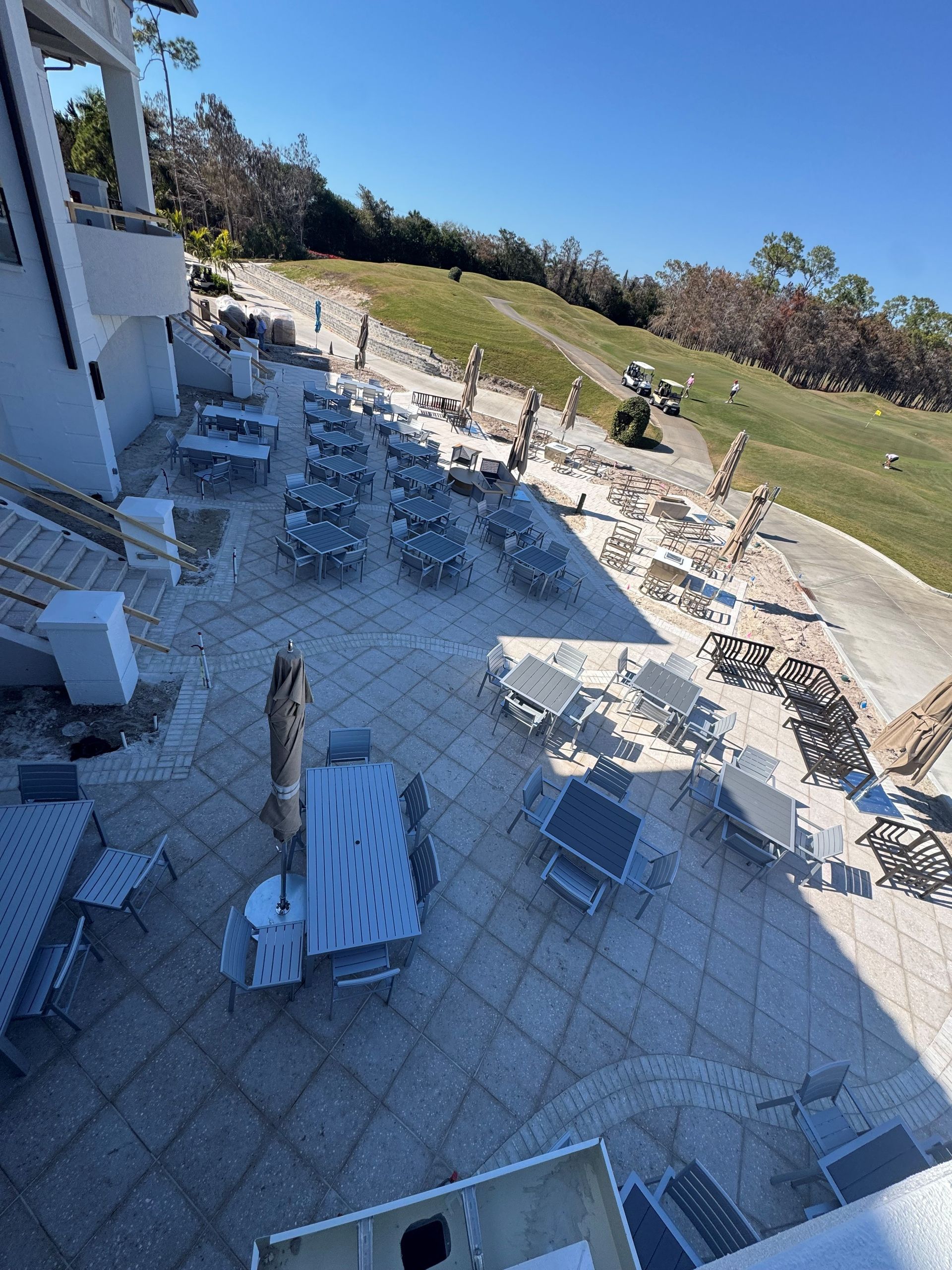 Empty outdoor patio with white tables and chairs beside a building, overlooking a grassy lawn under blue sky