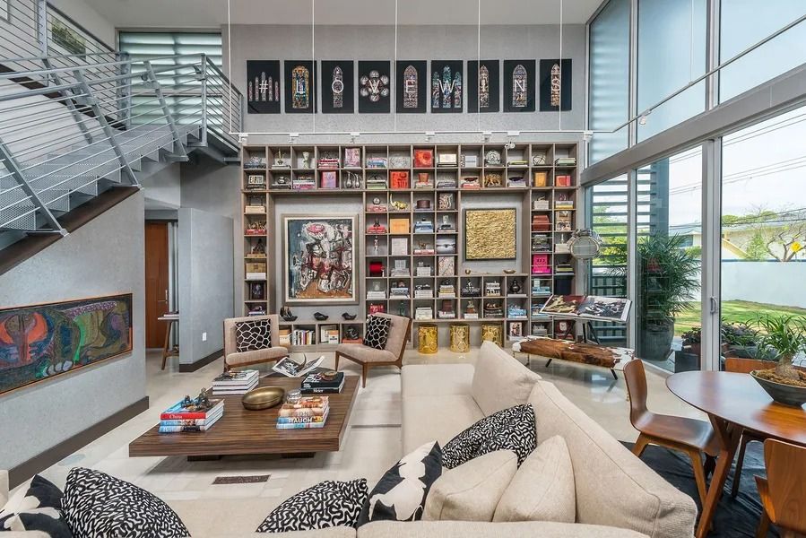 Modern living room with large bookshelf, artwork, and floor-to-ceiling windows; neutral tones.