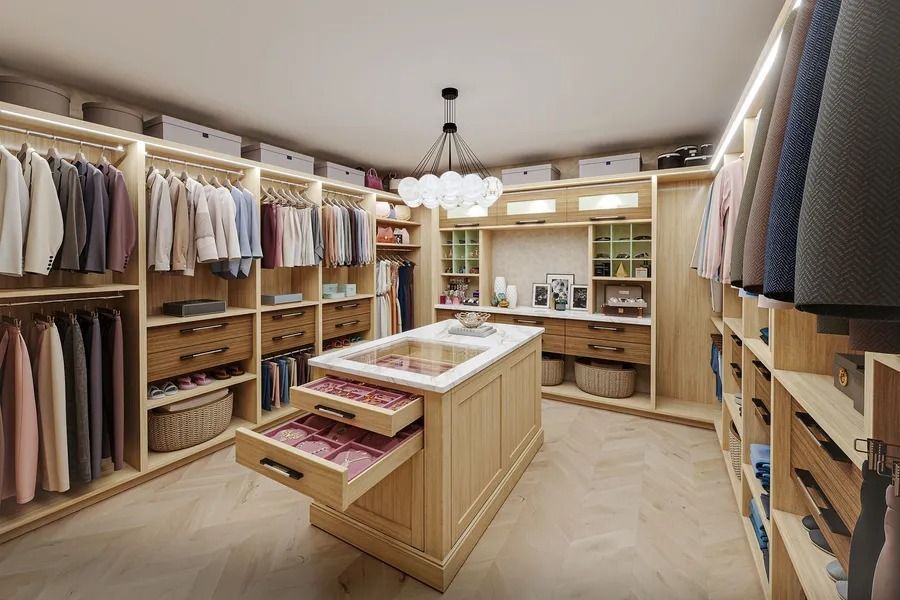 Luxurious walk-in closet with clothes, drawers, island with jewelry, and bright lighting.