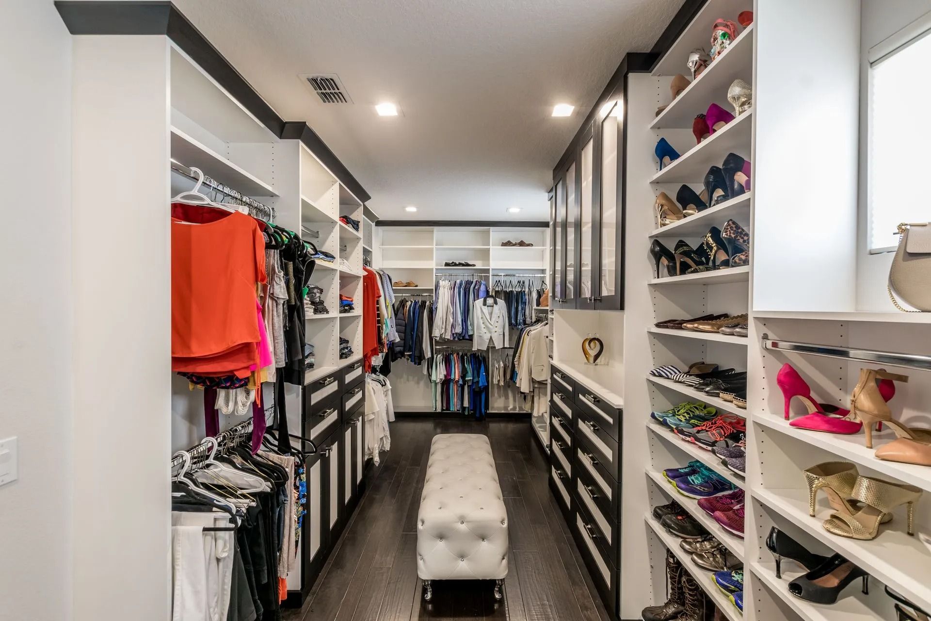 Walk-in closet with white shelving, black accents, and a tufted bench. Clothing and shoes are organized.