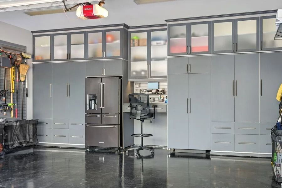 Gray garage storage cabinets with a refrigerator, desk, and cabinets with glass doors.