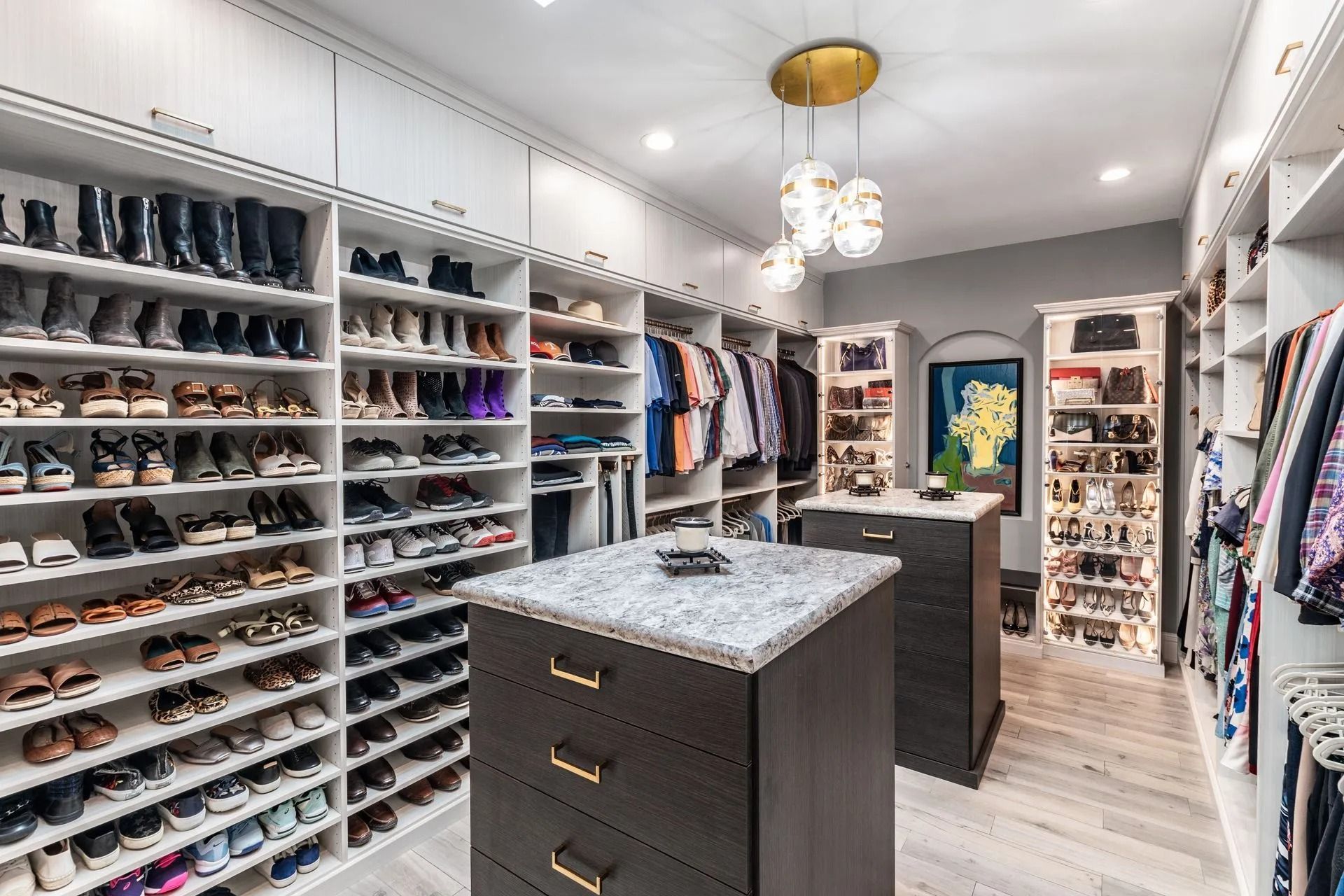 Walk-in closet with white shelves full of shoes and clothes, dark wood cabinets, and a decorative chandelier.
