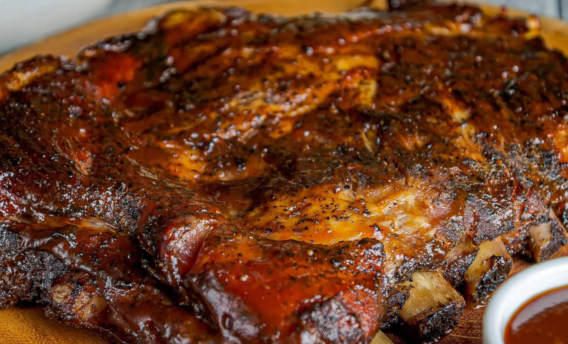 Rack of barbecue ribs, dark brown and glistening, served on a wooden platter with sauce.