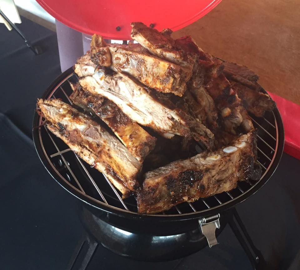 Pile of seasoned ribs on a charcoal grill, with a red lid in the background.
