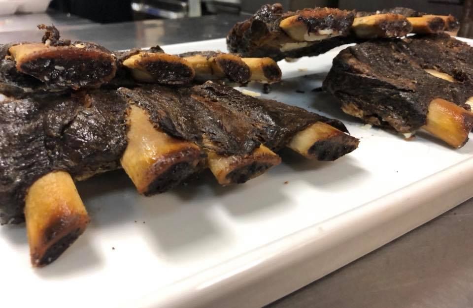 Smoked beef short ribs on a white cutting board, showing dark bark and exposed bones.