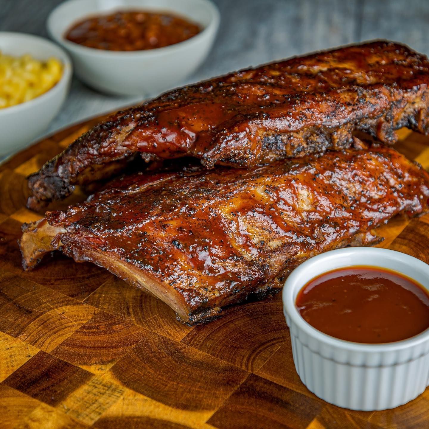 A wooden board holding two racks of glazed, dark-brown BBQ ribs, with a ramekin of sauce and side dishes in the background.