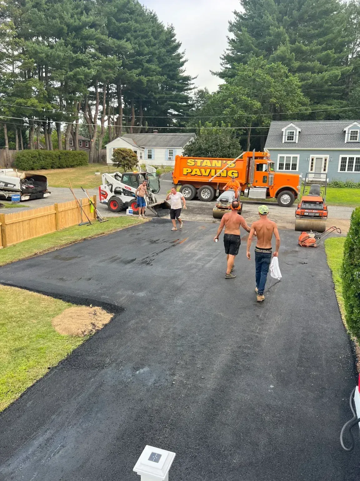 Workers paving a black driveway with orange trucks and equipment in a suburban neighborhood.