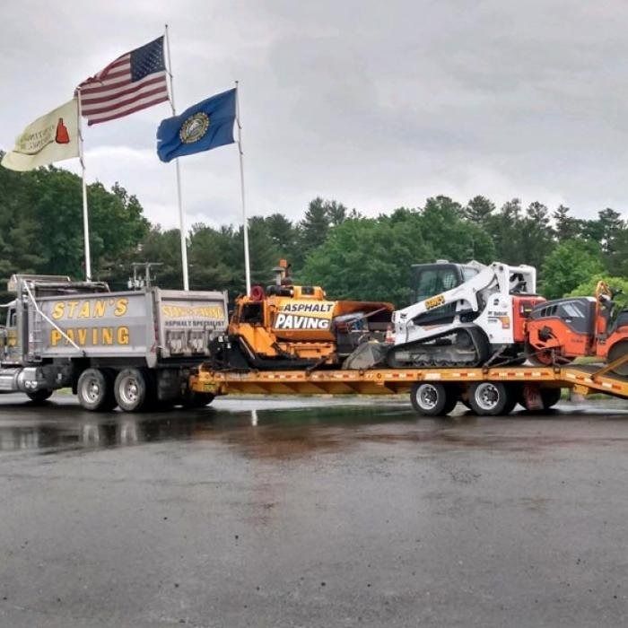 Tow truck hauling a flatbed with heavy equipment in a rainy lot, with American and state flags above.