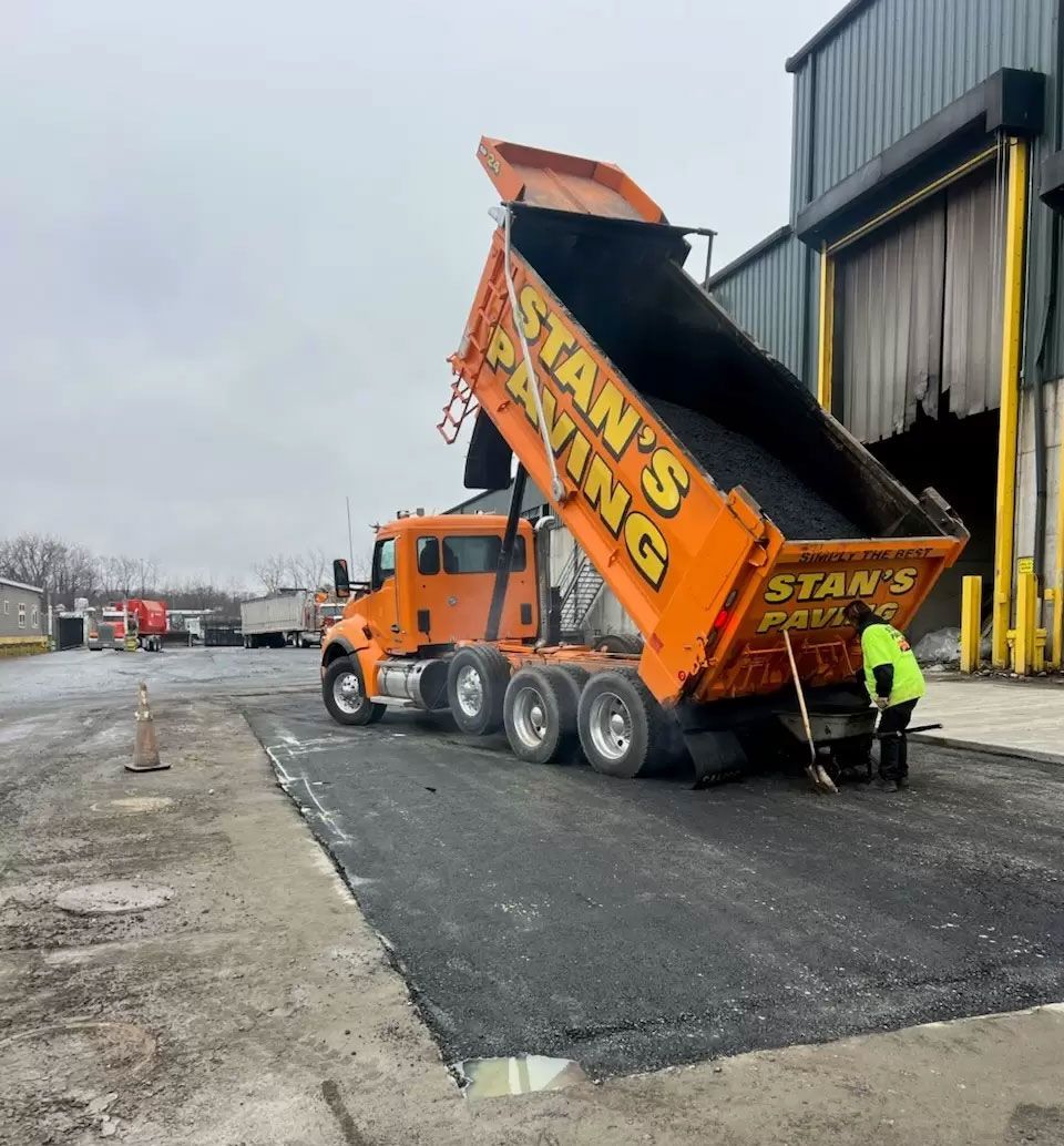Orange dump truck unloading gravel beside a warehouse on a wet road