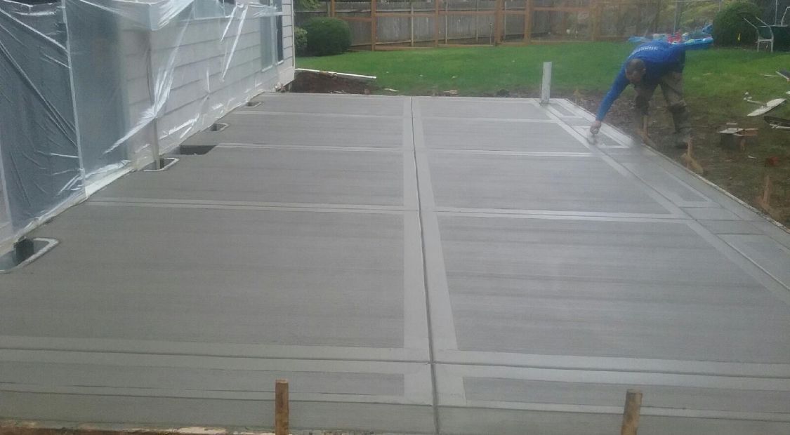 A concrete patio is being finished by a construction worker in a yard setting.