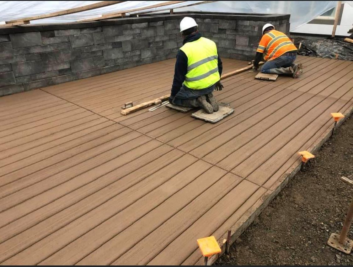 Construction workers laying brown deck boards. One kneels, using a level. A stone wall is in the background.