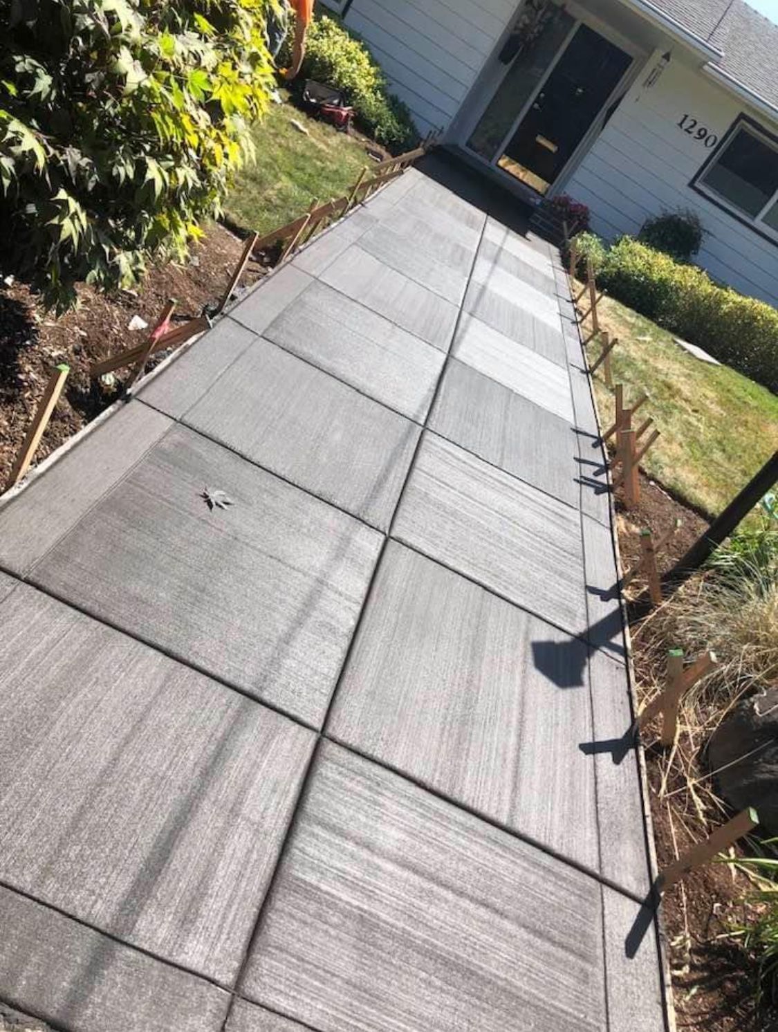 Gray tiled concrete walkway leading to a house, bordered by grass and wooden stakes.