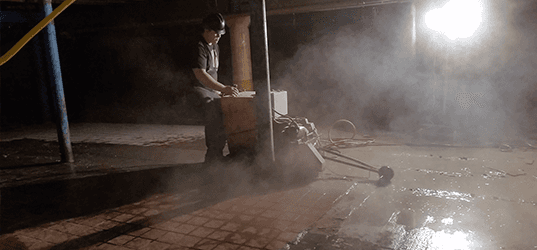 Worker pressure-washing a wet pavement at night beside a bright streetlight and mist