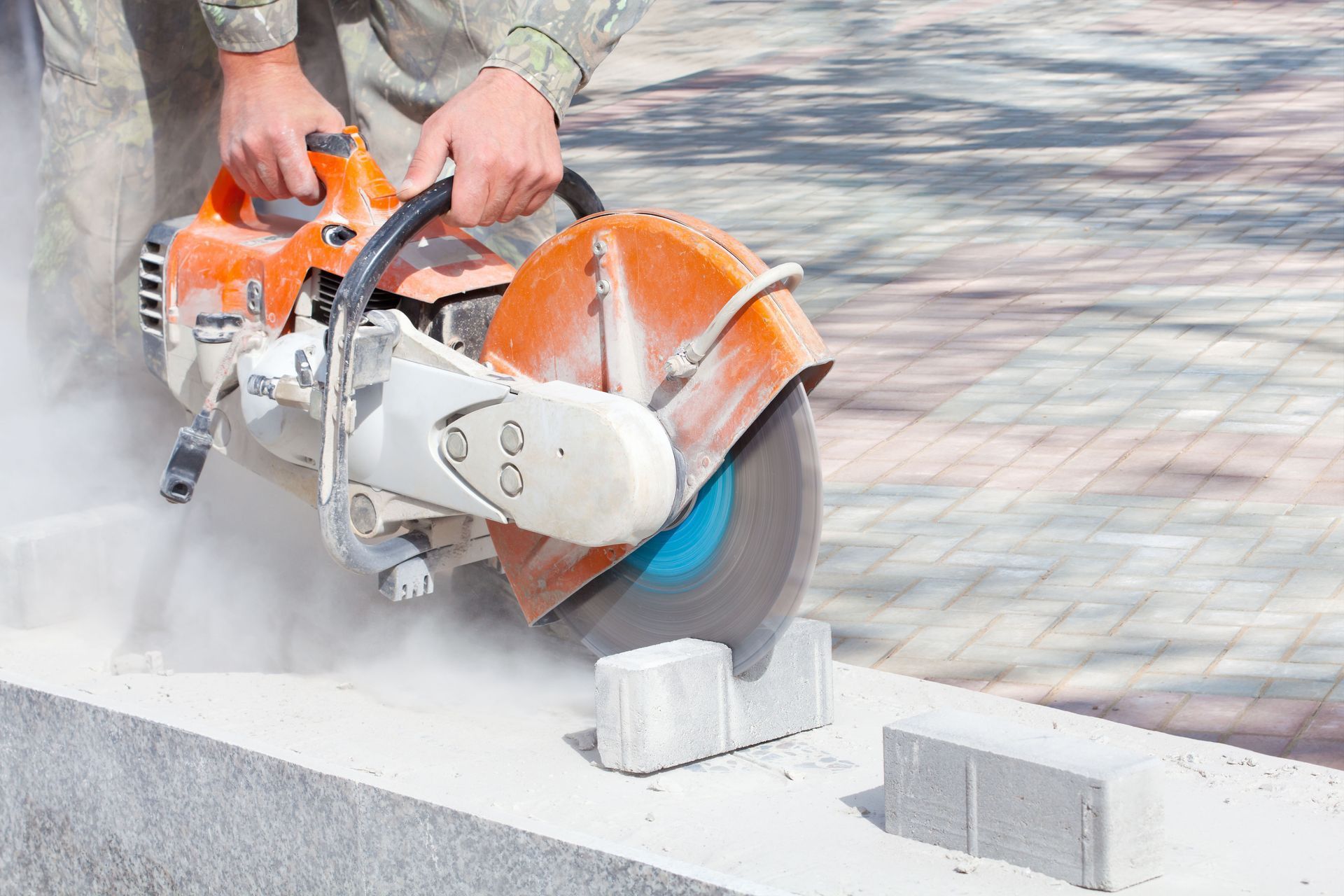 Worker using an orange concrete saw to cut paving stones, with dust on a sidewalk.