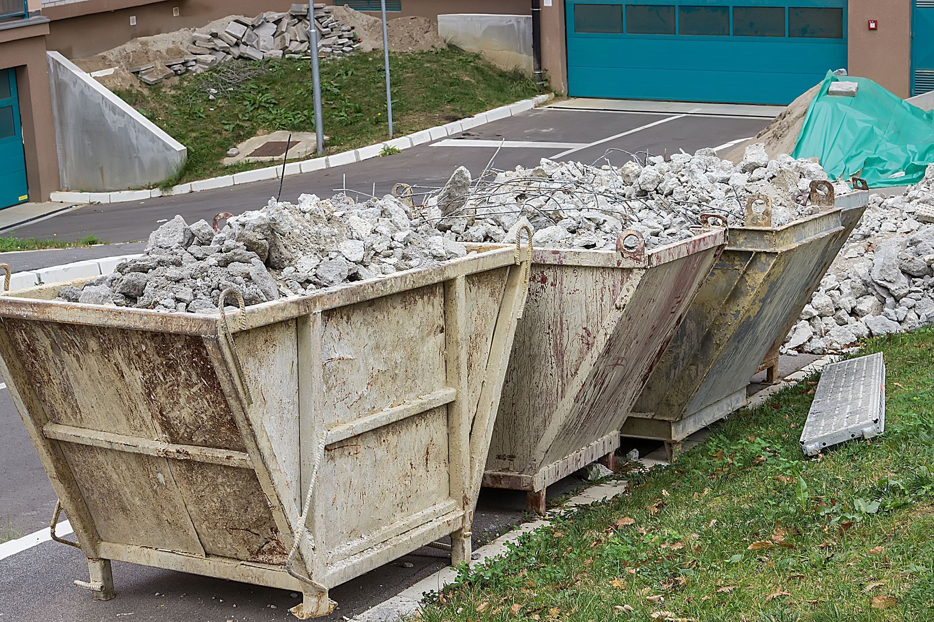 Two large metal dumpsters filled with rubble beside a paved road and grassy slope