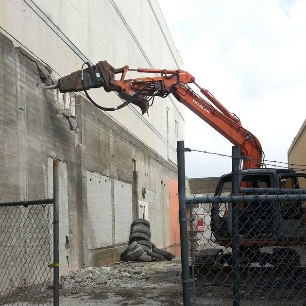 Orange excavator demolishing a concrete wall at a construction site behind a chain-link fence