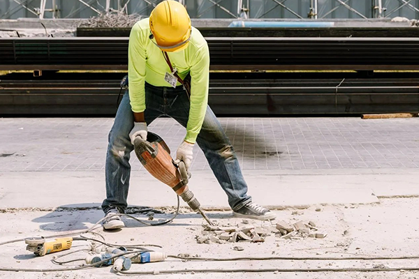 Worker in yellow hard hat using a jackhammer on a concrete surface at a construction site
