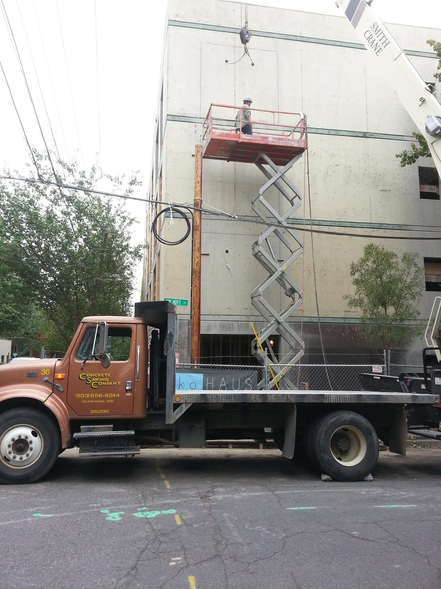 Brown utility truck with a raised scissor lift parked beside a building and trees.