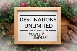 Logo for Destinations Unlimited travel agency, associated with Travel Leaders. Framed on a wood and greenery backdrop.