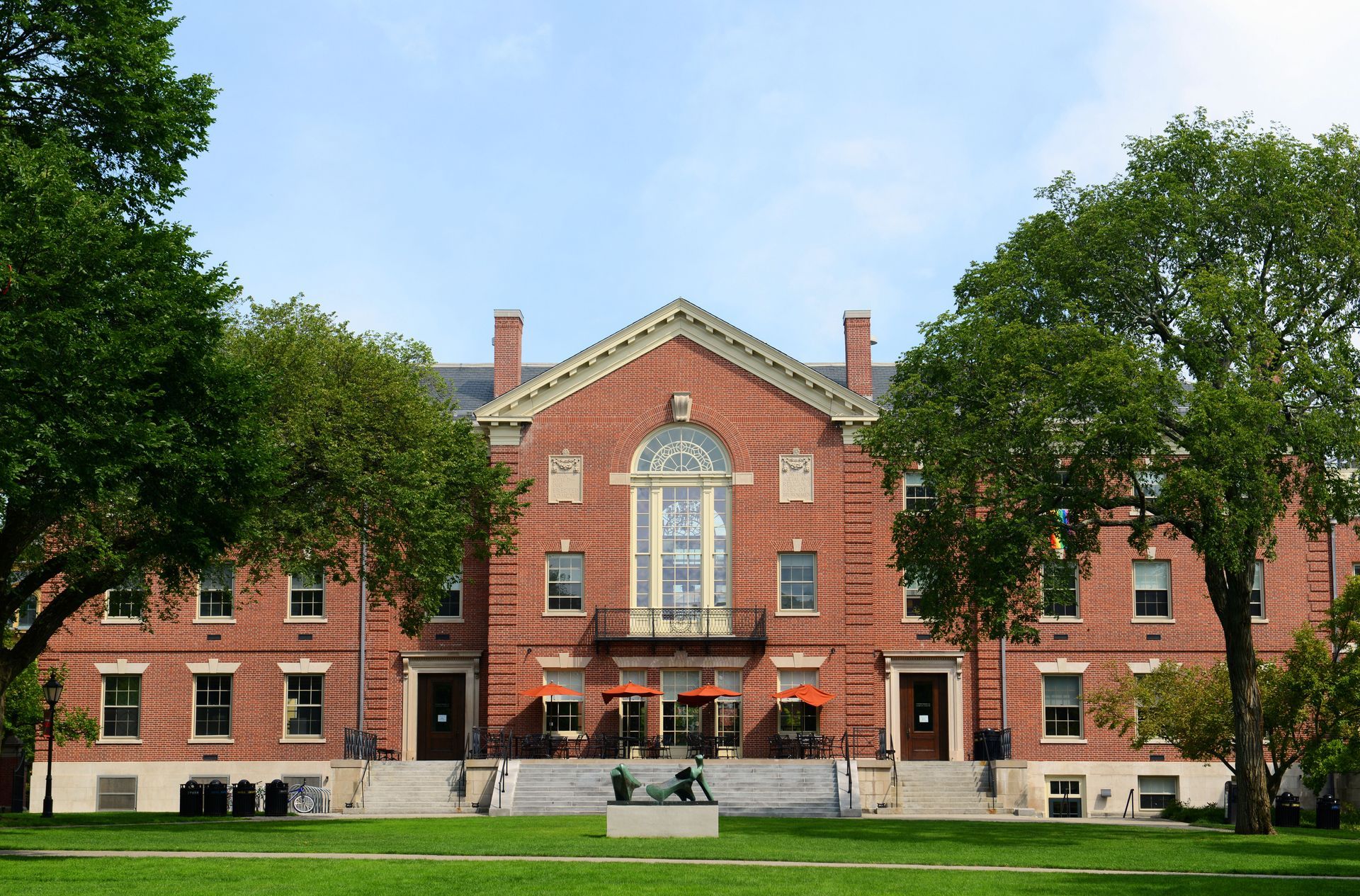 Red brick building with a pediment, balcony, and trees on a green lawn. Blue sky background.