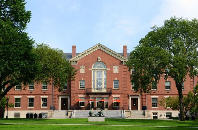 Red brick building with a pediment, balcony, and trees on a green lawn. Blue sky background.
