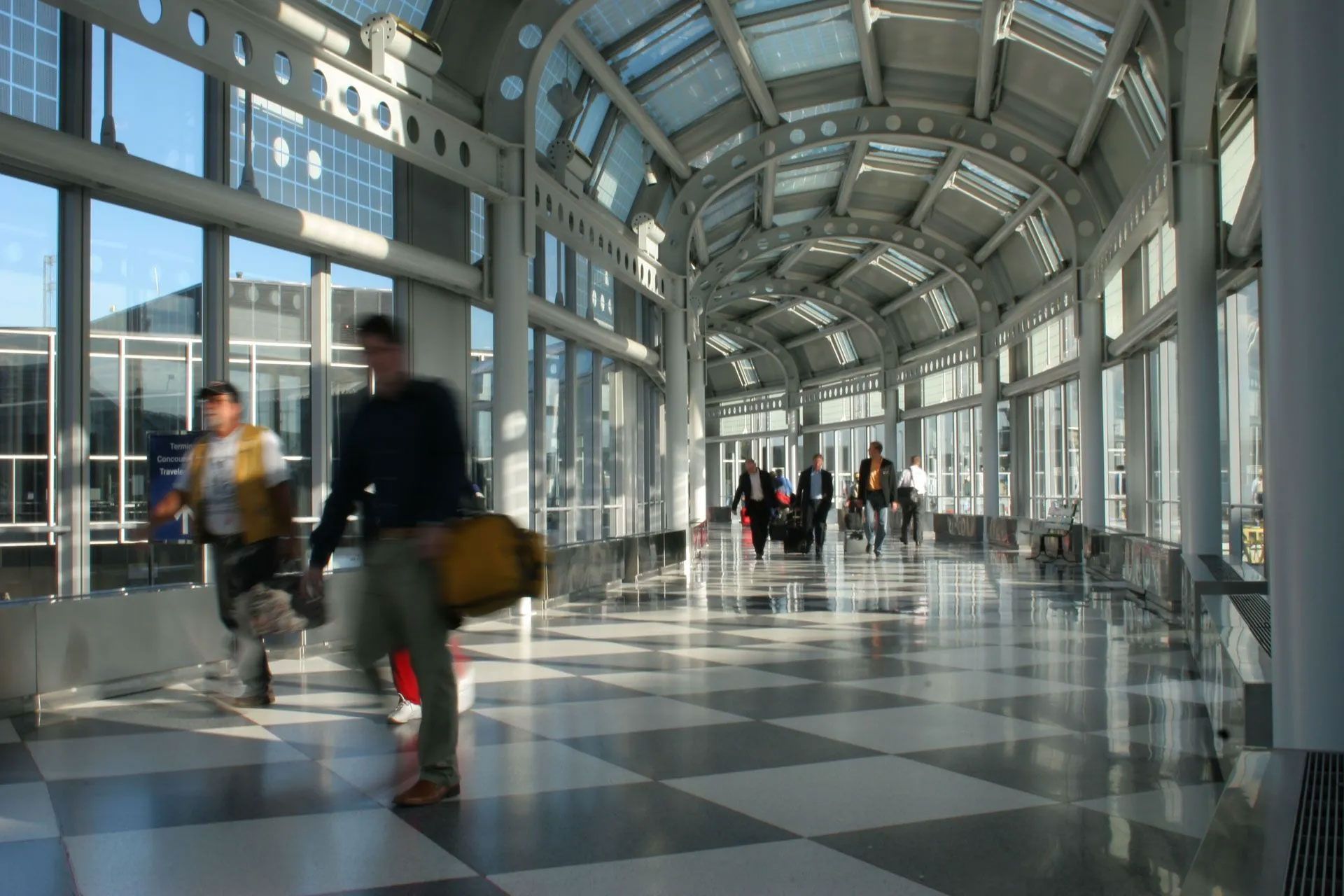 A sunlit airport walkway with a checkerboard floor, where people with luggage walk toward an exit.
