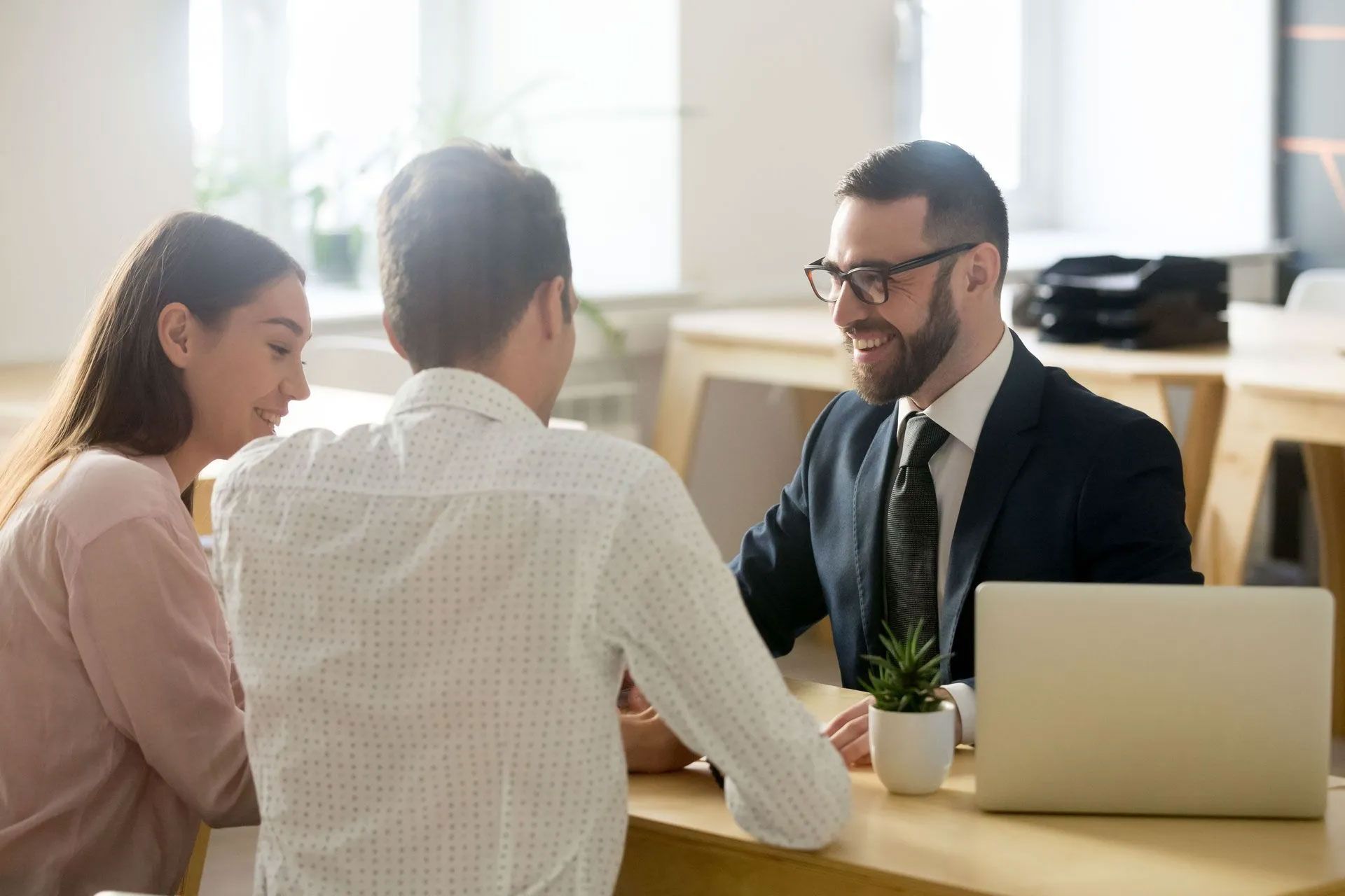 A professional meeting with a person in a suit advising two others sitting at a desk with a laptop.