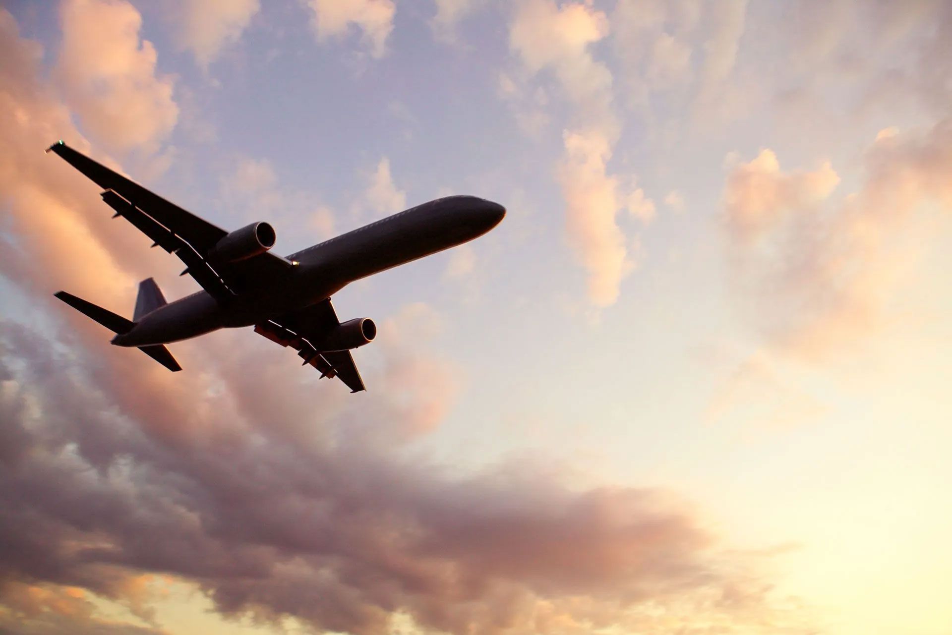 A silhouette of a passenger airplane flying upward against a sky filled with soft, golden-pink clouds at sunset.