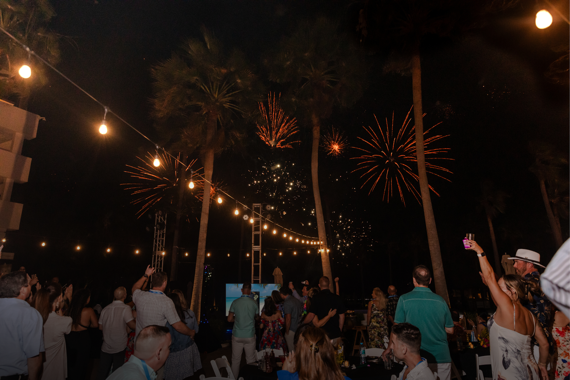 Outdoor nighttime crowd under string lights, watching fireworks burst in the sky.