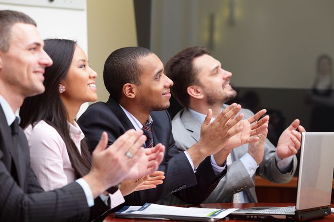 Four professionals in business attire applaud while looking toward a presentation off-screen.