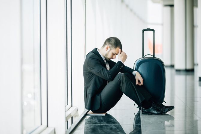 A person in a business suit sitting on the floor by a window in an airport terminal, looking distressed beside a suitcase.