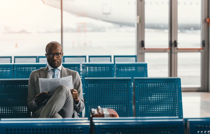 A person in a suit sits in an airport gate area reading a document, with a plane visible through the window behind them.