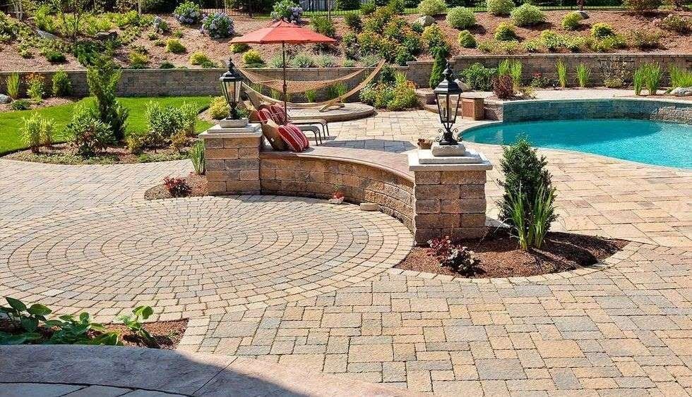 Patio with brick pavers, pool, retaining walls, hammock, and landscaping in sunny setting.