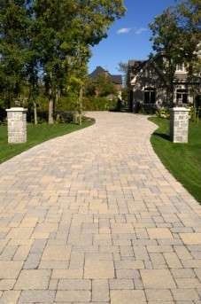 Brick paver driveway leading to a large house, flanked by green grass and stone pillars.