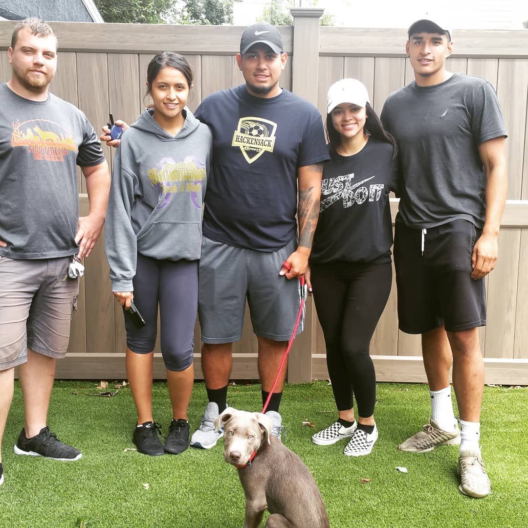 Five people stand together on a lawn in front of a wooden fence, accompanied by a small grey puppy on a red leash.