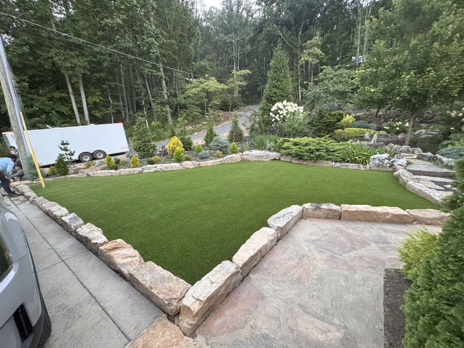 A small, fenced yard with green turf, stone edging, and landscaping. A vehicle is on the left.
