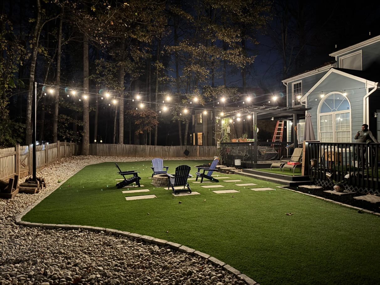 Nighttime backyard with string lights, fire pit area, chairs on artificial grass, and a house.