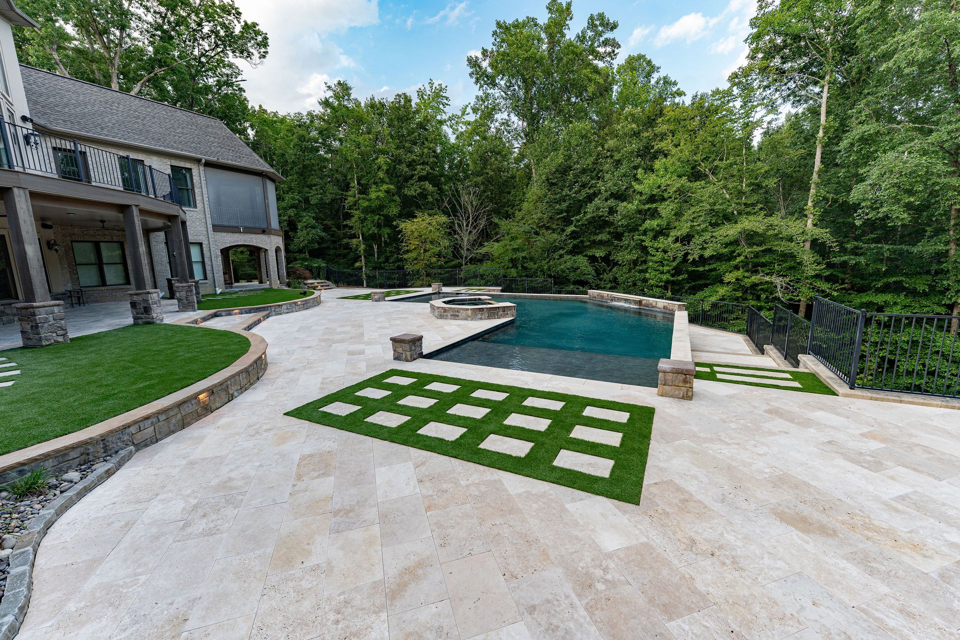 Stone patio with pool and house in wooded setting. Green lawn and square grass insets.