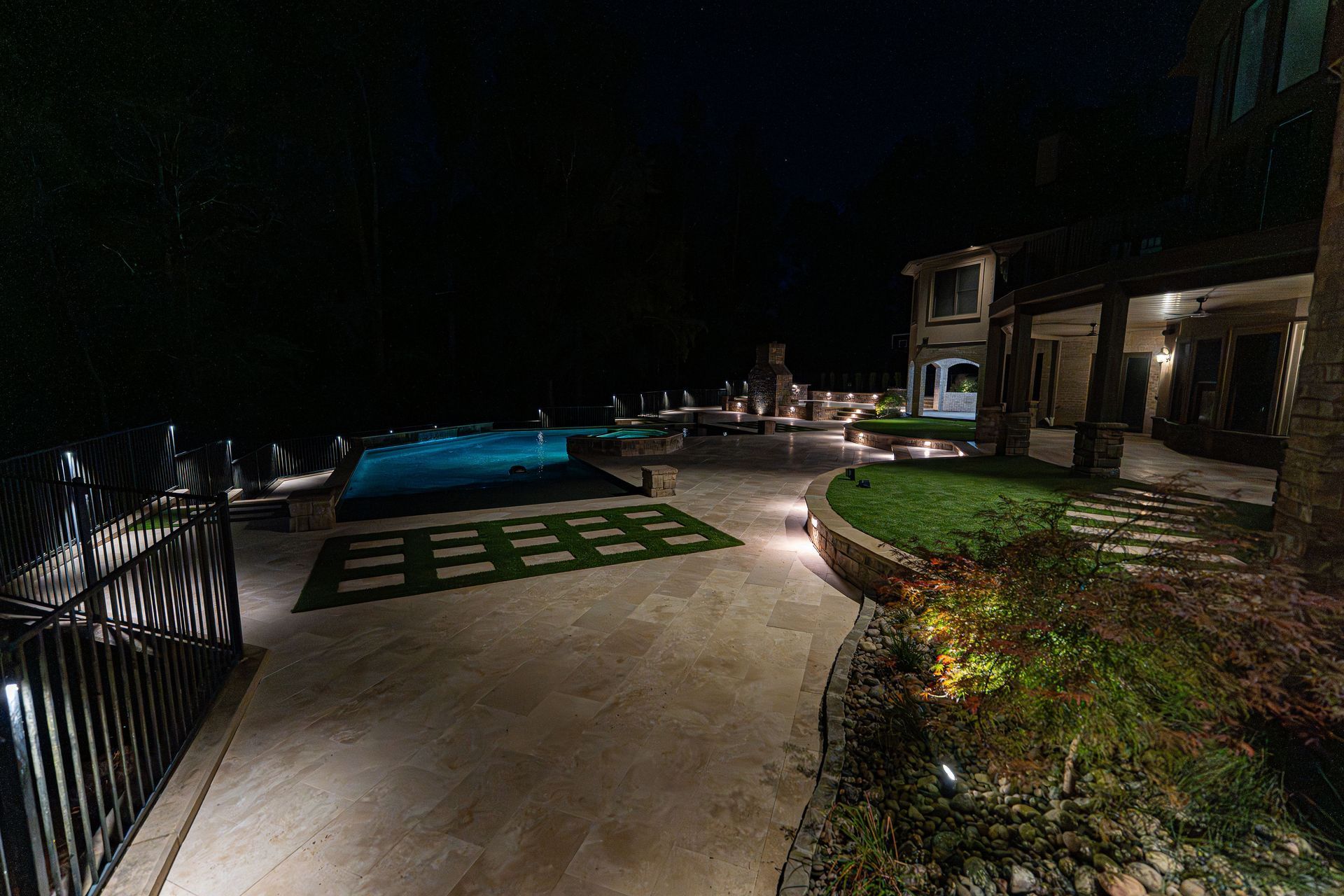 Night view of a luxury backyard with a pool, landscaping, and a house illuminated by spotlights.