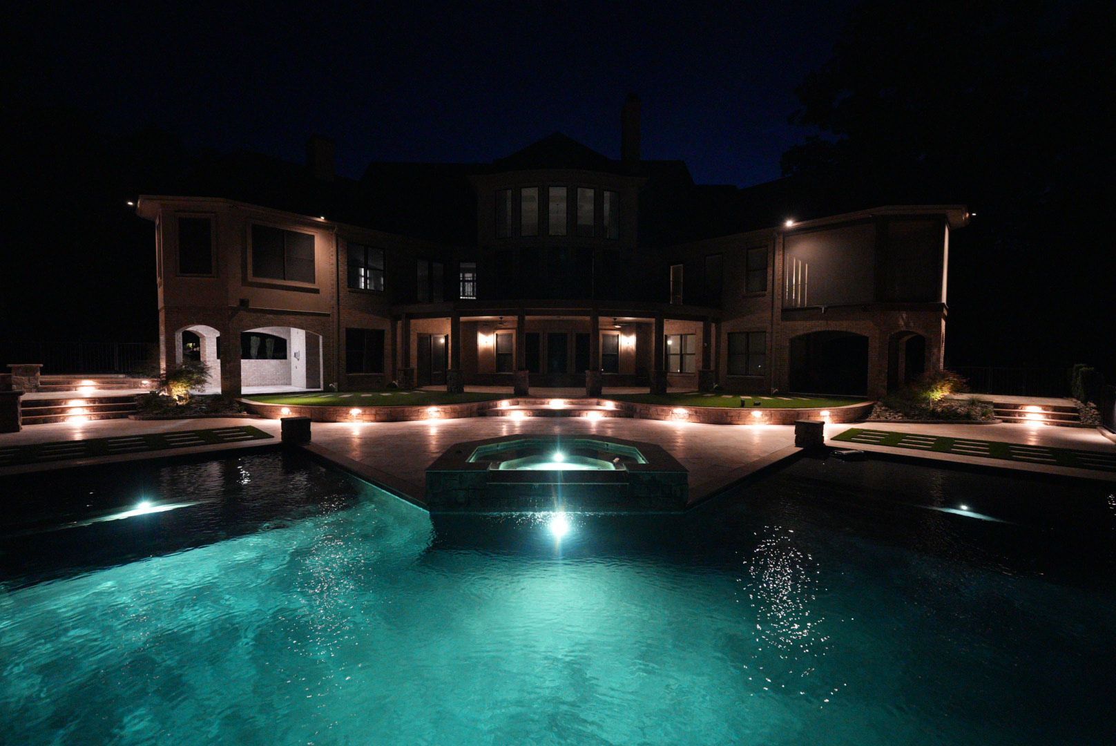 Luxury mansion illuminated at night with a pool, spa, and lights; dark sky.