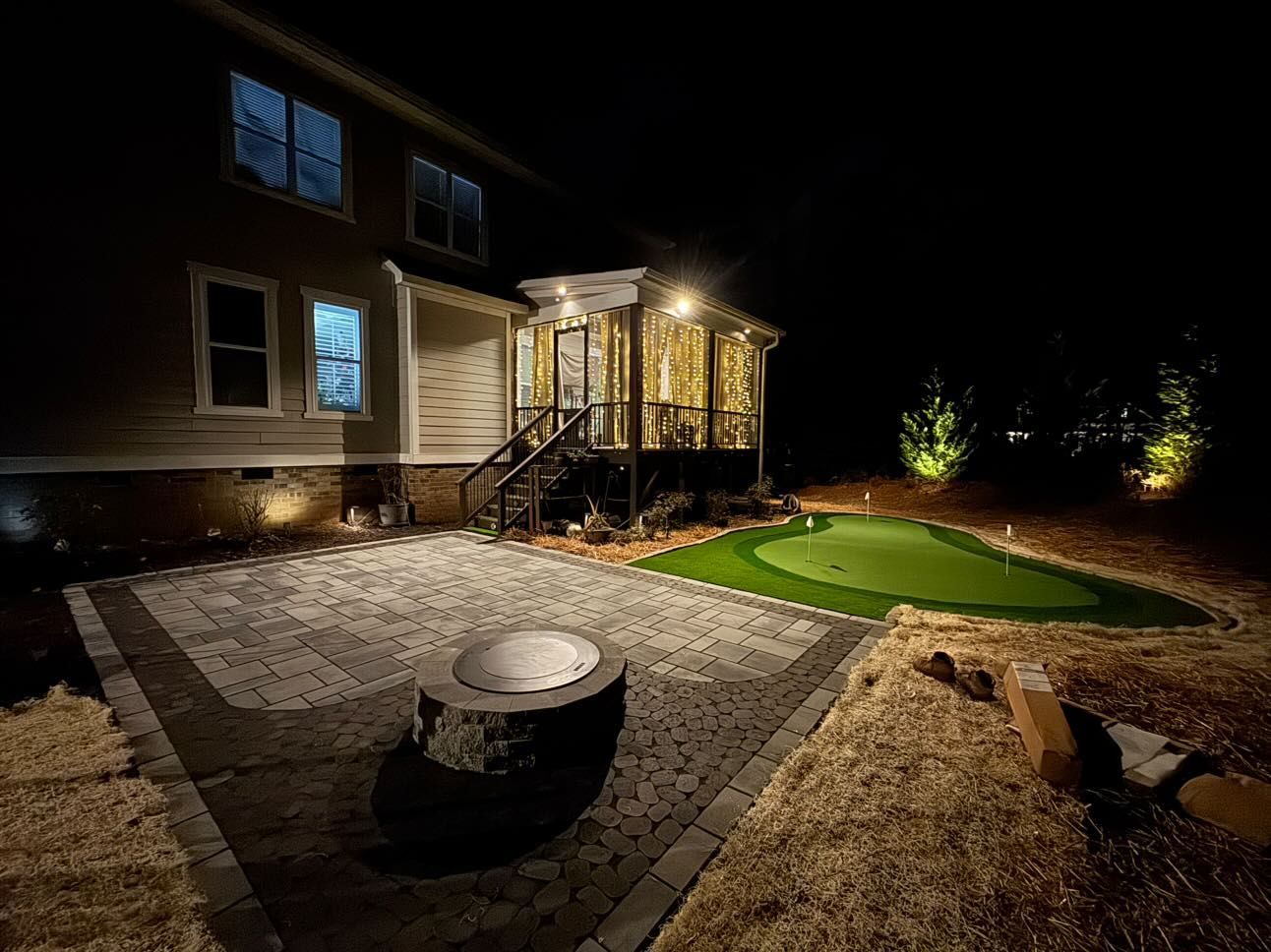 Nighttime backyard scene with a patio, fire pit, and a putting green, all lit by landscape lights. The house is visible in the background.
