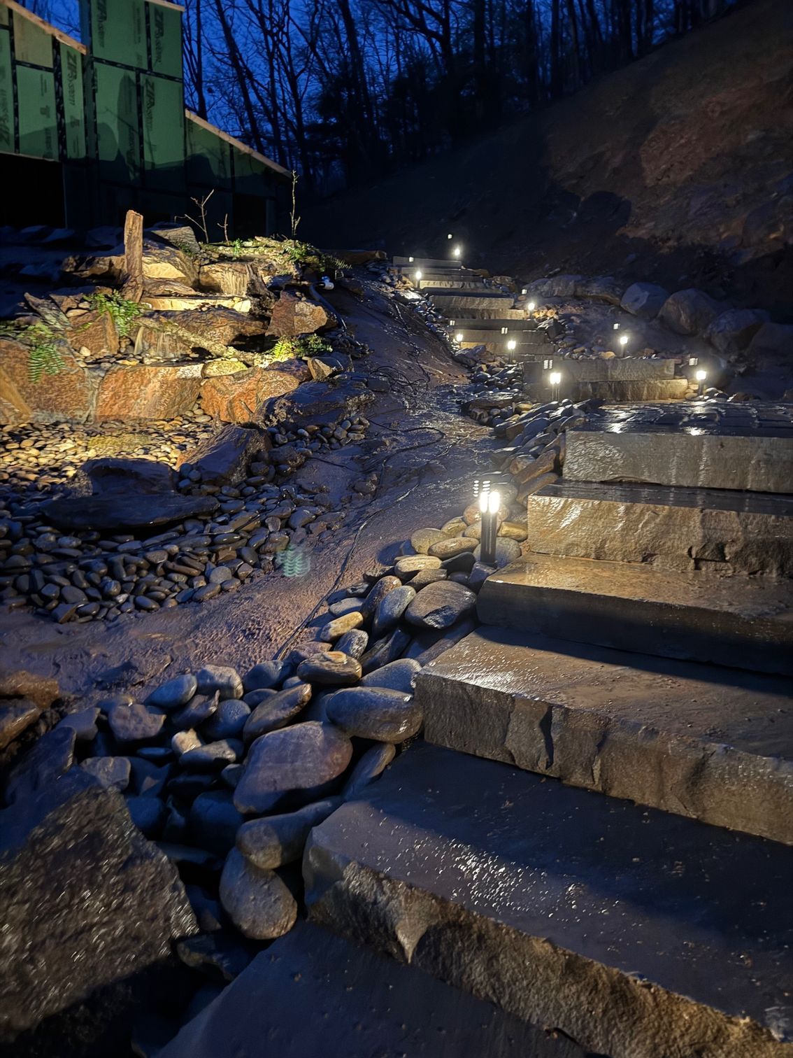 Stone steps with pathway lights lead uphill, beside a rocky embankment, lit up at night.