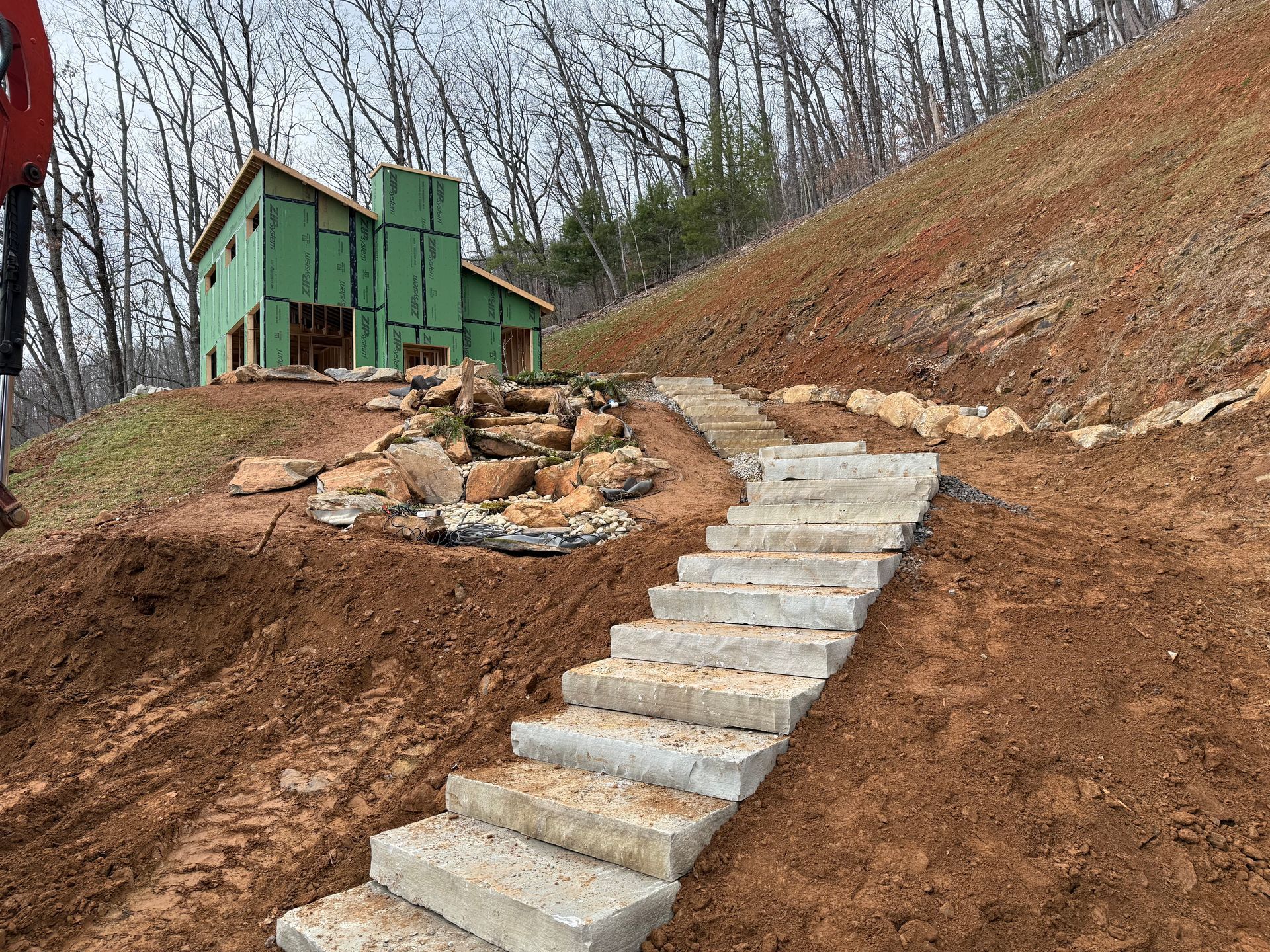 Concrete steps ascending a hillside towards a house under construction, with green sheathing. Brown dirt surrounds the steps and building.