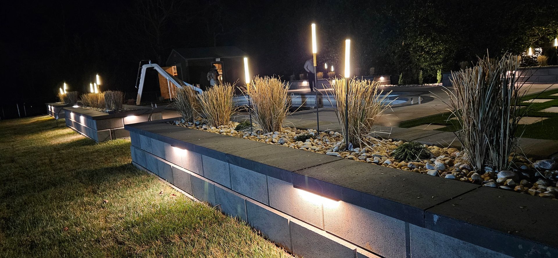 Nighttime view of a landscaped yard with low-lit retaining walls, ornamental grasses, and vertical light poles.