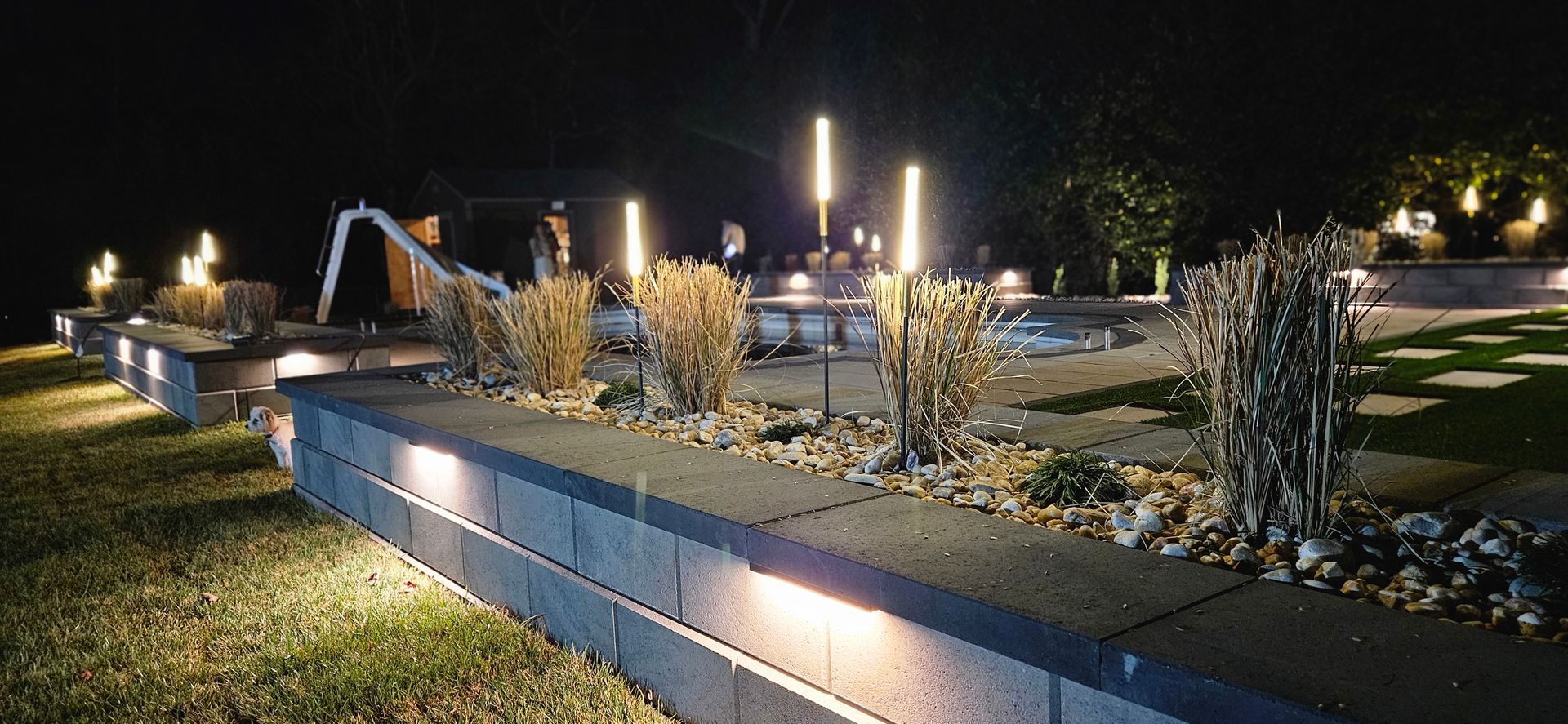 Night view of a landscaped garden with built-in lighting illuminating a retaining wall and various plants. Tall vertical lights glow in the background.