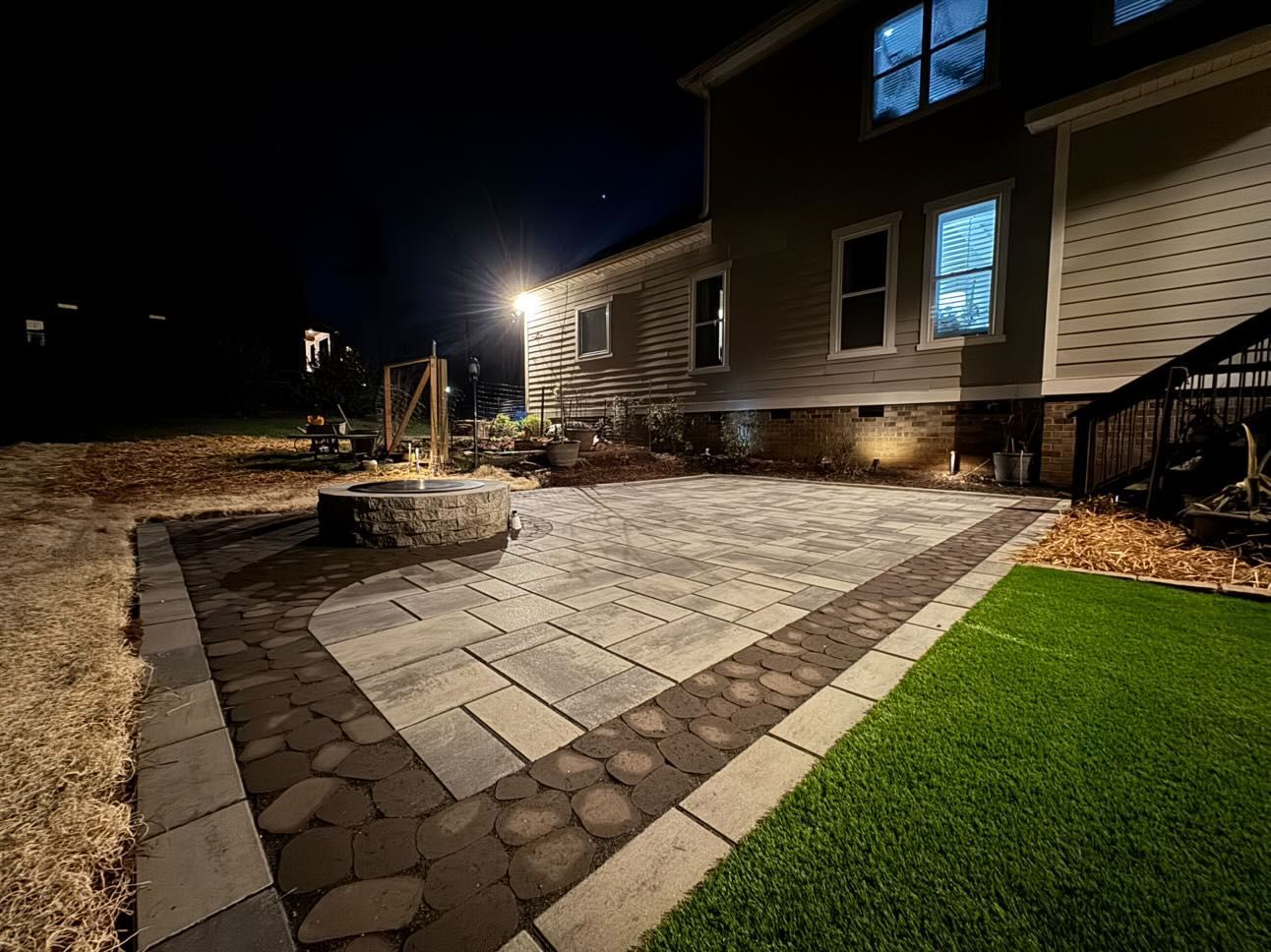 A backyard patio at night with a fire pit, pavers, and a house in the background. Green turf and mulch surround the patio.