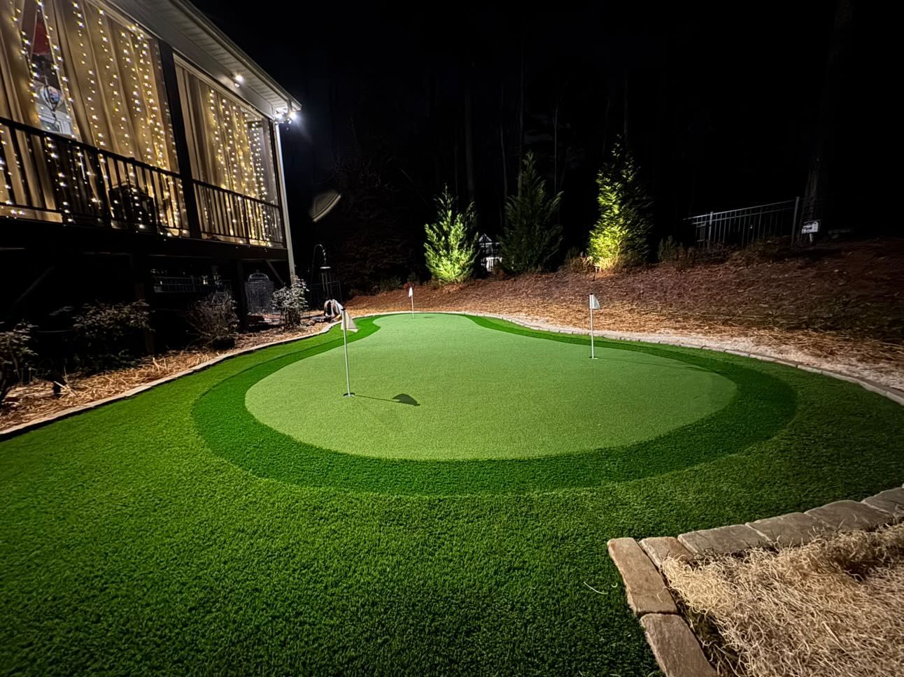 A backyard putting green at night, illuminated by spotlights. The green features multiple tiers and flags.