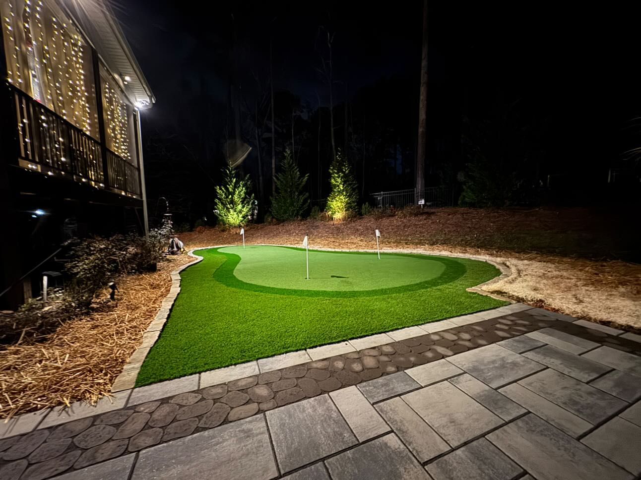A nighttime view of a backyard putting green, brightly lit, surrounded by pavers, mulch, and trees.